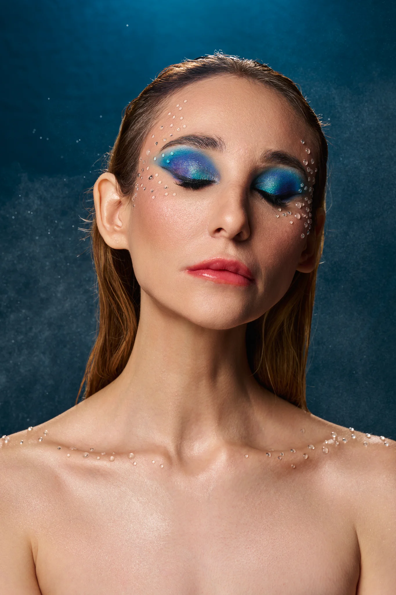 Beauty portrait with blue editorial makeup and crystal accents, water mist visible in background, studio photography