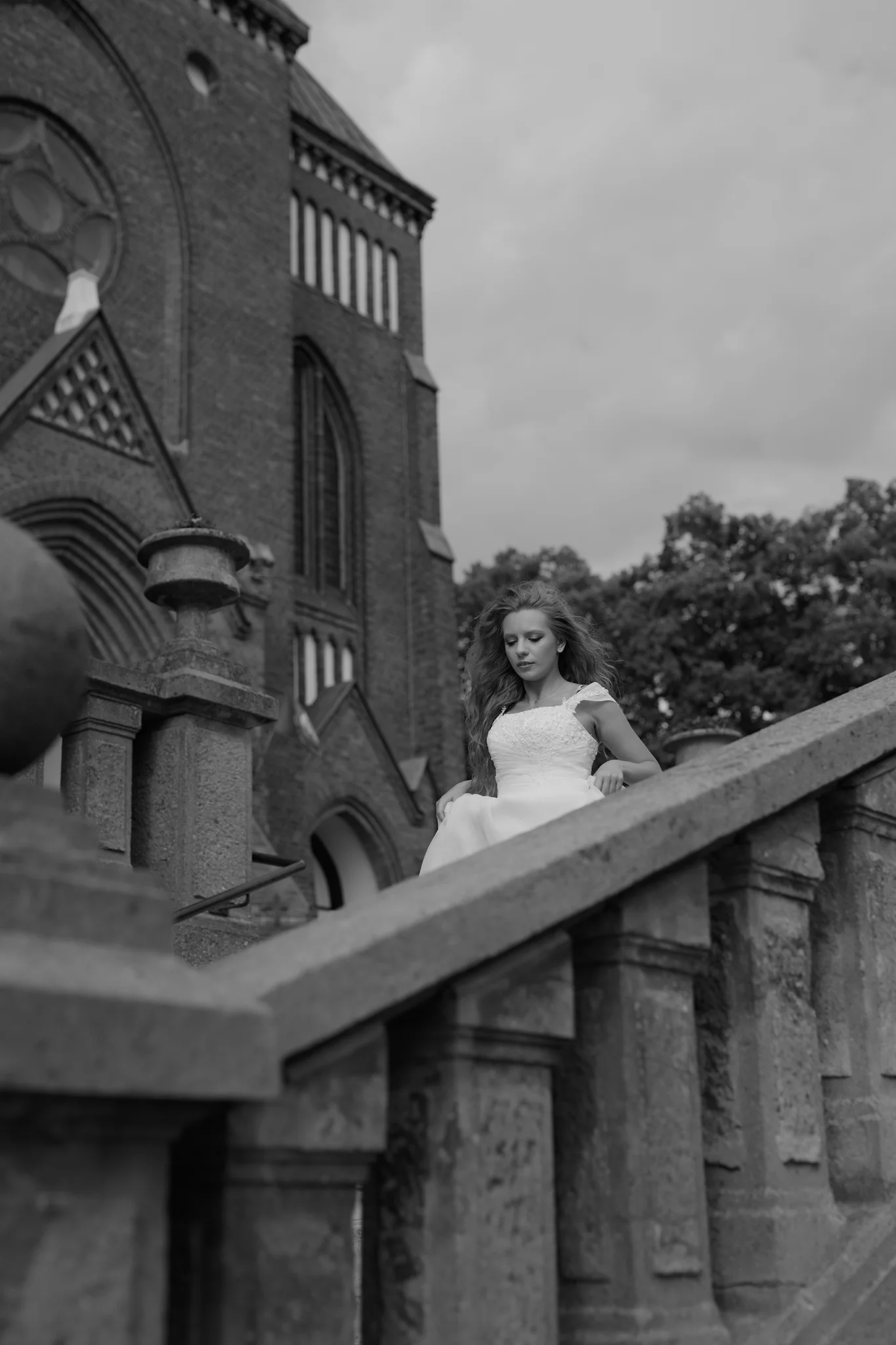 Woman in wedding dress on stone steps beside a Gothic church, hair caught in the wind, low-angle shot, black and white outdoor portrait.