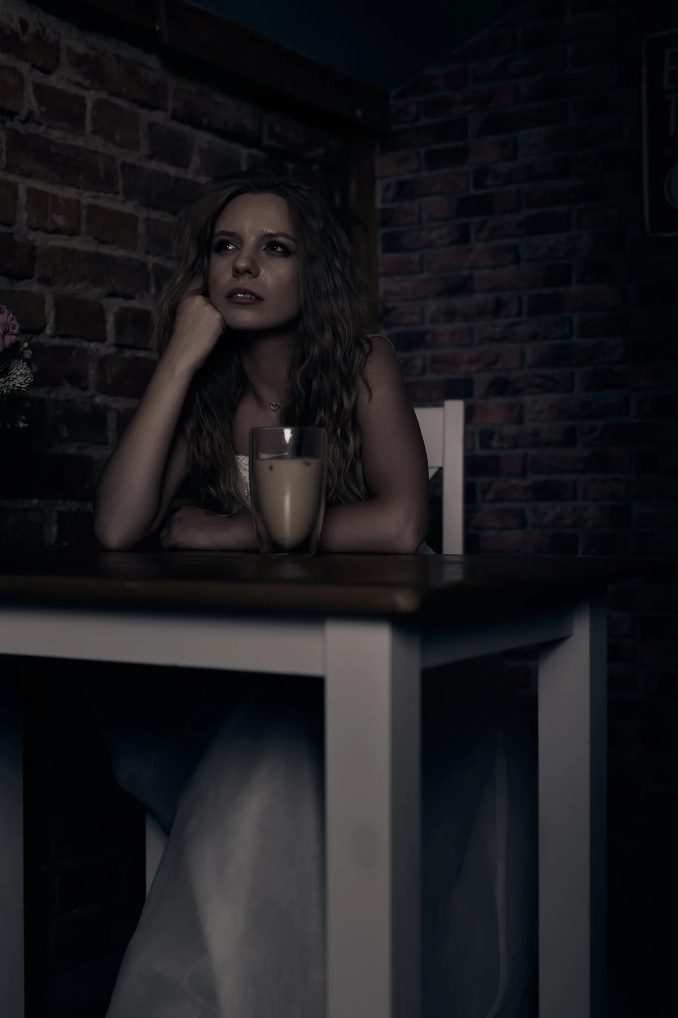 Woman in wedding dress sitting at a table in a dark café with brick wall background, head resting on hand, pensive expression, glass of coffee on the table.