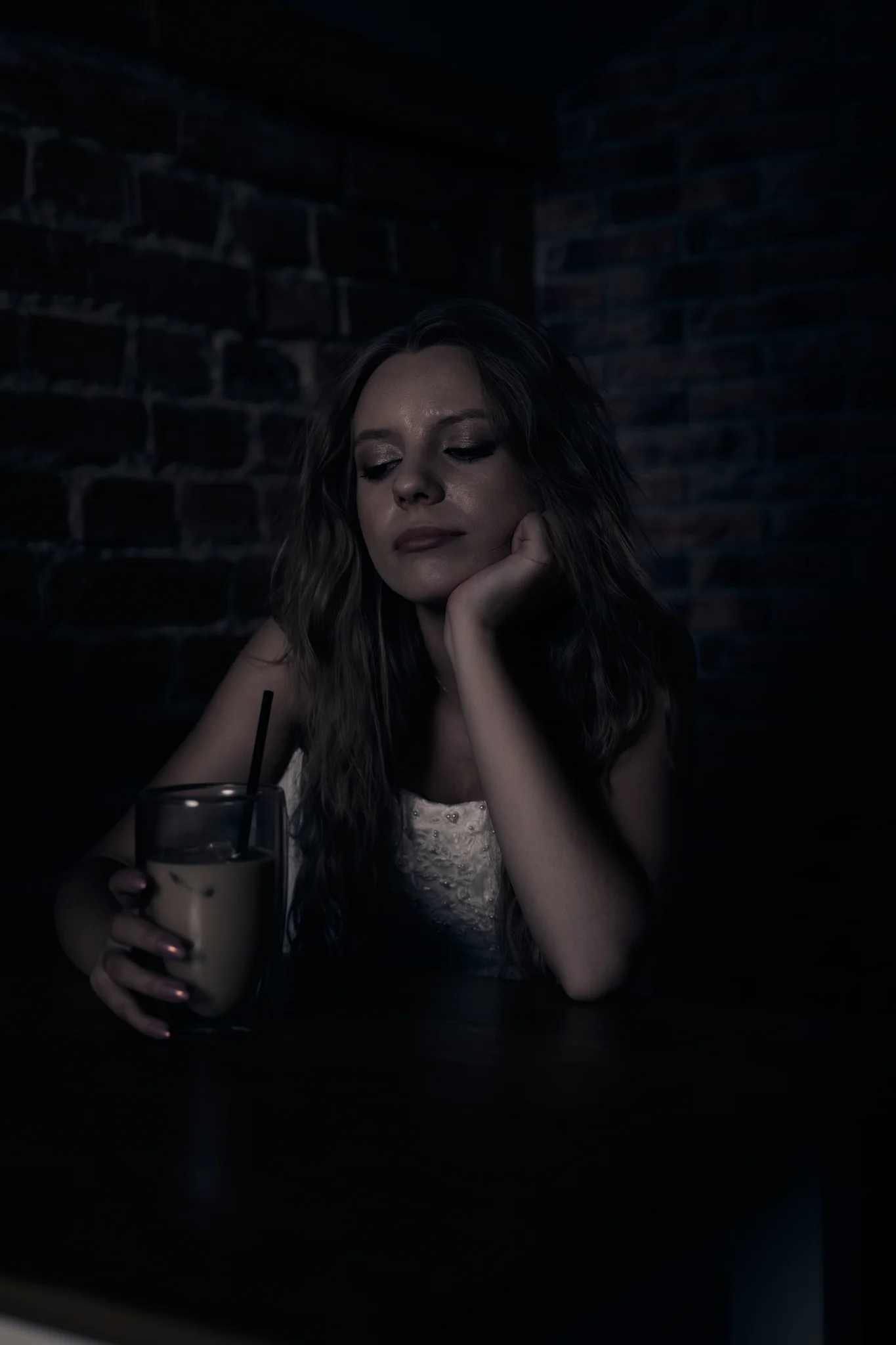 Portrait of woman in wedding dress in a dark café, eyes closed, head resting on hand, holding a glass of coffee.