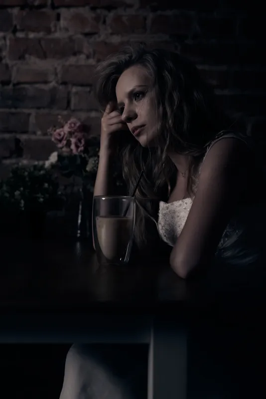 Woman in wedding dress at a table in a dark café, gaze turned to the side, flowers in the background, glass of coffee on the table.
