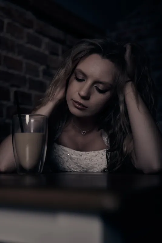 Woman in wedding dress in a dark café, head resting on both hands, eyes closed, glass of coffee in the foreground.